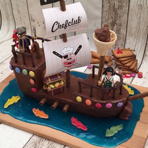 Gâteau d'anniversaire en forme de bateau pirate, avec un glaçage au chocolat, des gâteaux roulés pour les canons et des voiles en papier avec des têtes de mort.