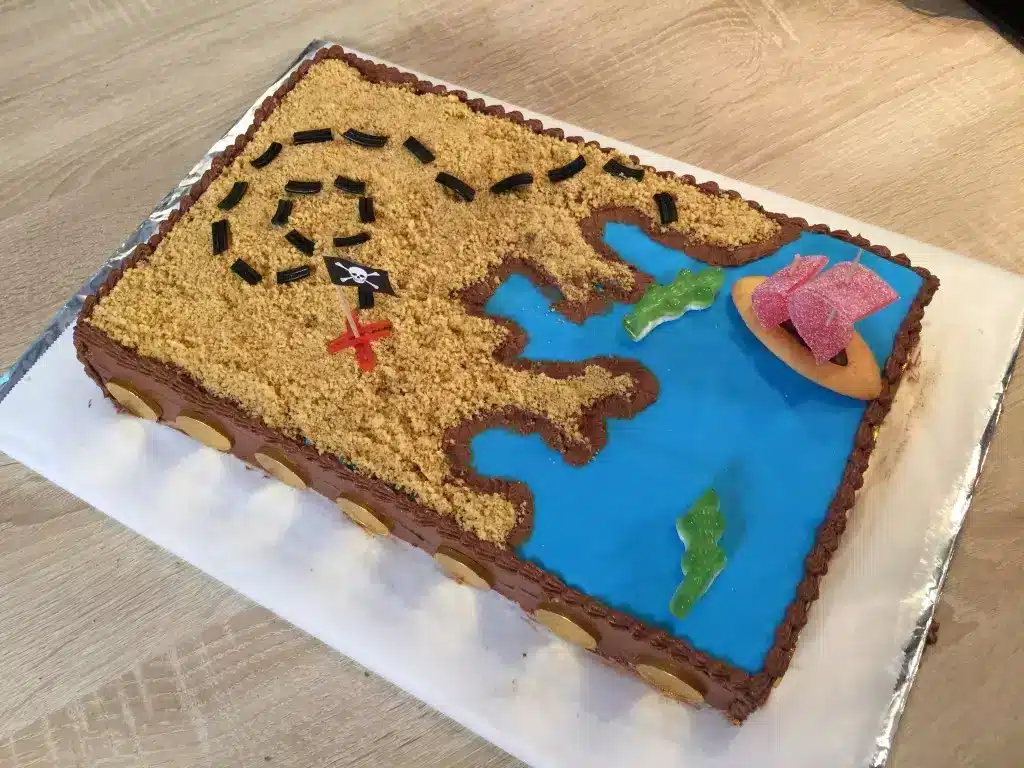 Gâteau d'anniversaire rectangulaire décoré comme une carte au trésor. Une moitié est bleue avec des poissons en bonbon pour la mer, l'autre est couverte de miettes de biscuits pour le sable avec un chemin en réglisse menant à une croix rouge.